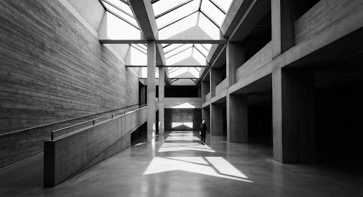 A striking high-contrast black and white architectural portrait of a modern, brutalist museum space. Sunlight streams through geometric skylights, casting dramatic, sharp shadows across the polished concrete floor. A solitary figure stands in the distance, emphasizing scale. The mood is contemplative, intellectual, and sophisticated, aligning perfectly with a high-end minimalist editorial aesthetic.