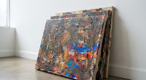 A detail shot of highly textured, layered oil paintings stacked against a stark white gallery wall. The camera focuses on the thick impasto strokes revealing brilliant flashes of cobalt blue and crimson beneath darker, earthy overpainting. The minimalist gallery setting contrasts sharply with the chaotic energy of the canvases, creating a compelling intellectual tension.