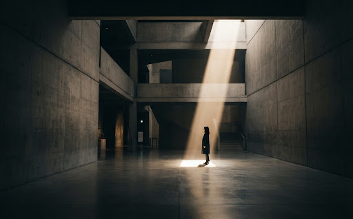 A wide, stunning photograph of a vast, minimalist concrete architectural space with a single beam of dramatic sunlight cutting through the darkness, illuminating a lone figure. High contrast, warm stone and deep shadow tones, reflecting a quiet, contemplative mood.