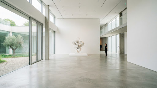 A wide architectural shot of a minimalist gallery interior in bright natural light. The space features clean lines, white walls, and a single prominent sculpture, creating a calm, intellectual atmosphere characteristic of modern editorial photography.