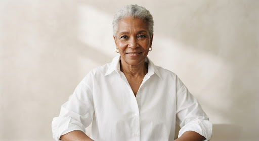 A documentary-style portrait of an older Black woman with short silver hair, wearing an oversized crisp white shirt, looking directly at the camera with a confident expression. Photographed against an ivory background with soft shadow. High-end editorial aesthetic, minimalist, sharp focus on the face.