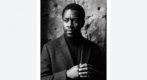 A stark documentary portrait of a Black author against a textured stone wall, wearing a tailored coat. The lighting is dramatic with strong shadows, creating a serious tone. High-end editorial aesthetic, sharp focus, monochromatic feel.
