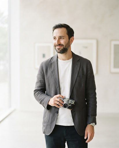 A minimalist portrait of a male photographer. He holds a vintage film camera slightly lowered. The aesthetic is clean, light-mode, with a focus on subtle textures and a very shallow depth of field. The image feels candid yet perfectly composed, fitting for a contributor bio in a luxury art and culture magazine.
