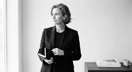 A sophisticated black and white portrait of an intellectual magazine editor. The subject is looking thoughtfully off-camera, set against a pristine, minimalist white background. Soft, diffused natural light highlights the subtle textures of their tailored linen jacket. The aesthetic is extremely high-end, calm, and evocative of premium editorial portraiture found in luxury lifestyle journals.