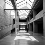 A striking high-contrast black and white architectural portrait of a modern, brutalist museum space. Sunlight streams through geometric skylights, casting dramatic, sharp shadows across the polished concrete floor. A solitary figure stands in the distance, emphasizing scale. The mood is contemplative, intellectual, and sophisticated, aligning perfectly with a high-end minimalist editorial aesthetic.
