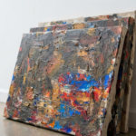 A detail shot of highly textured, layered oil paintings stacked against a stark white gallery wall. The camera focuses on the thick impasto strokes revealing brilliant flashes of cobalt blue and crimson beneath darker, earthy overpainting. The minimalist gallery setting contrasts sharply with the chaotic energy of the canvases, creating a compelling intellectual tension.