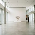 A wide architectural shot of a minimalist gallery interior in bright natural light. The space features clean lines, white walls, and a single prominent sculpture, creating a calm, intellectual atmosphere characteristic of modern editorial photography.