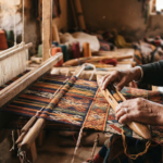 A refined, close-up photograph of artisan hands carefully weaving a vibrant, complex textile pattern. The lighting is warm, directional natural light spilling from a side window, highlighting the texture of the threads and the dust motes in the air. The color palette focuses on rich earth tones, golds, and deep indigo. The aesthetic feels tactile, culturally rich, and meticulously documented.