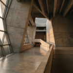 A wide, sweeping architectural shot of a modern, brutalist museum interior, bathed in warm, golden-hour light streaming through massive geometric windows. The composition is highly asymmetric, featuring vast negative space that highlights the stark concrete textures and the solitary figure of a person observing an art piece in the distance. The mood is contemplative, intellectual, and deeply rooted in high-end editorial photography.